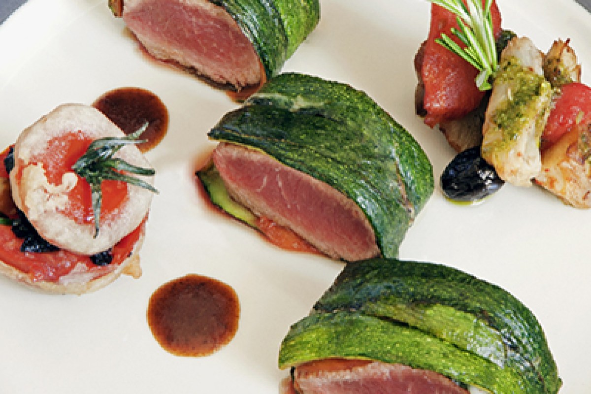 Plated dish of seared pink meat wrapped in greens with tomato, olive and shrimp garnish