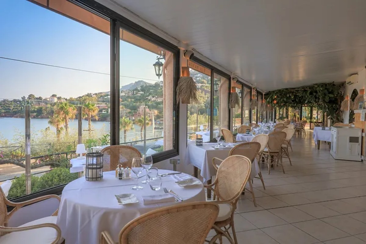 Restaurant terrace dining room with white-clothed tables, wicker chairs, and floor-to-ceiling windows overlooking the sea and coastline