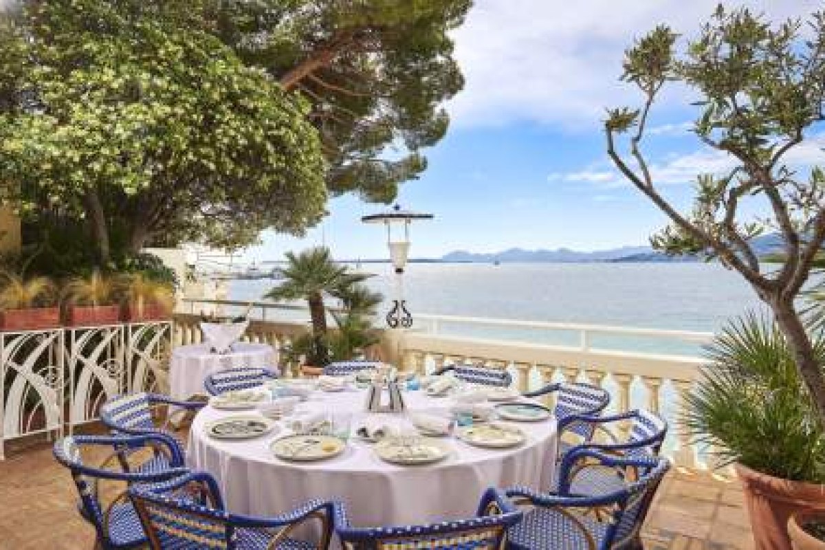 Terrace dining table set with blue wicker chairs overlooking the sea, framed by trees and a lantern