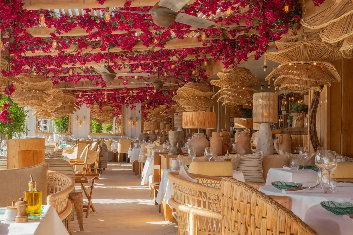 The dining room at La Petite Plage in Saint-Tropez, with a bougainvillea-covered pergola ceiling, woven rattan lamps, wicker chairs and a sand floor