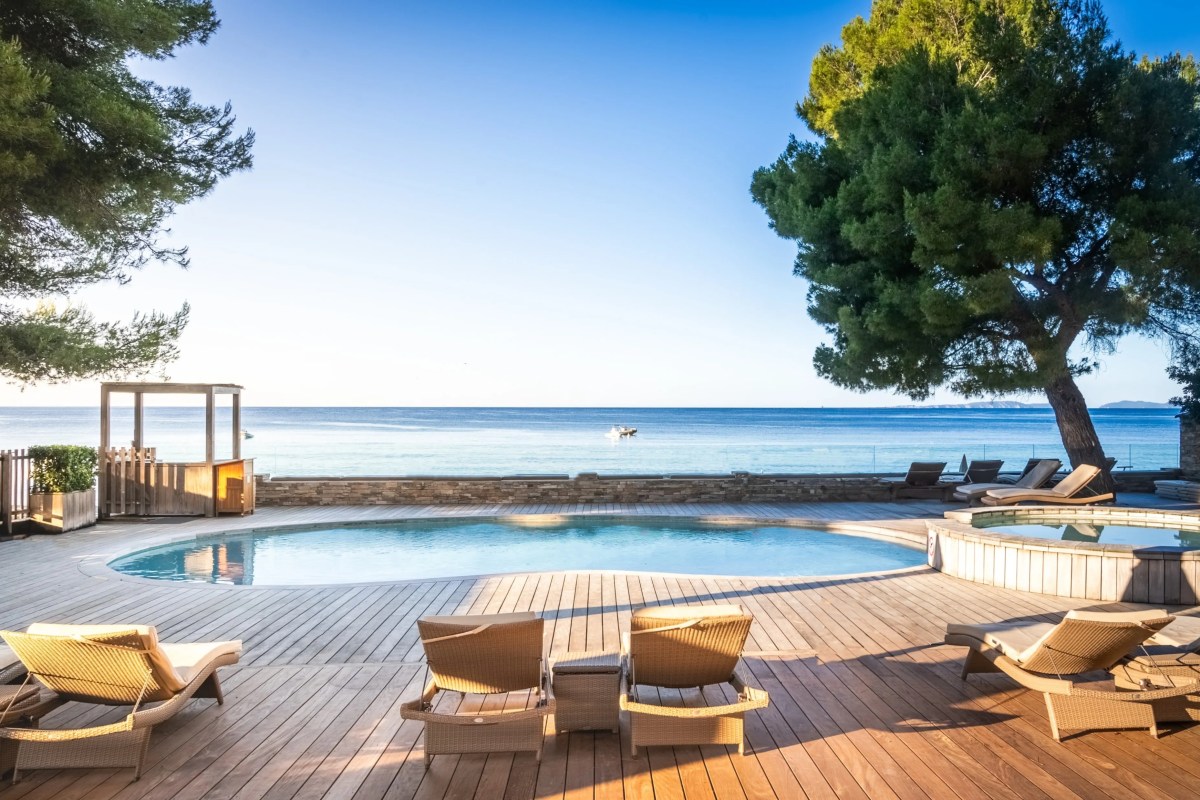 Outdoor pool deck with wooden sunloungers, a hot tub, and a view of the sea between trees