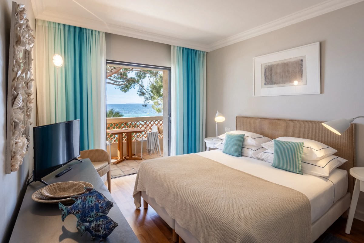 Hotel bedroom with a king bed, teal curtains, and a balcony opening to a sea view through pine trees