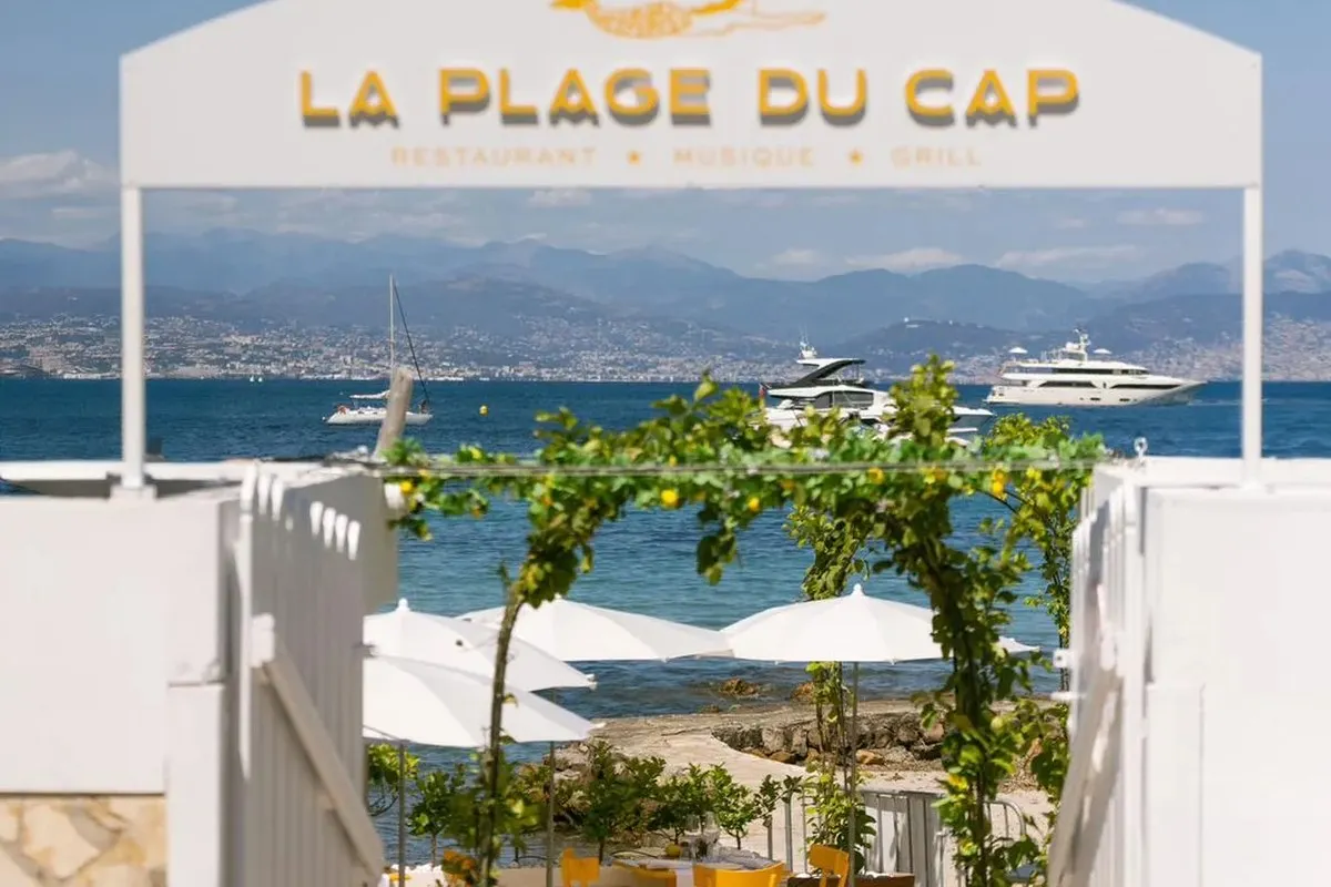 The entrance arch of La Plage du Cap on the Garoupe, with its sign «La Plage du Cap – Restaurant, Musique, Grill», a lemon-tree arch, white parasols and yachts on the bay, with the Baie des Anges and the mountains behind