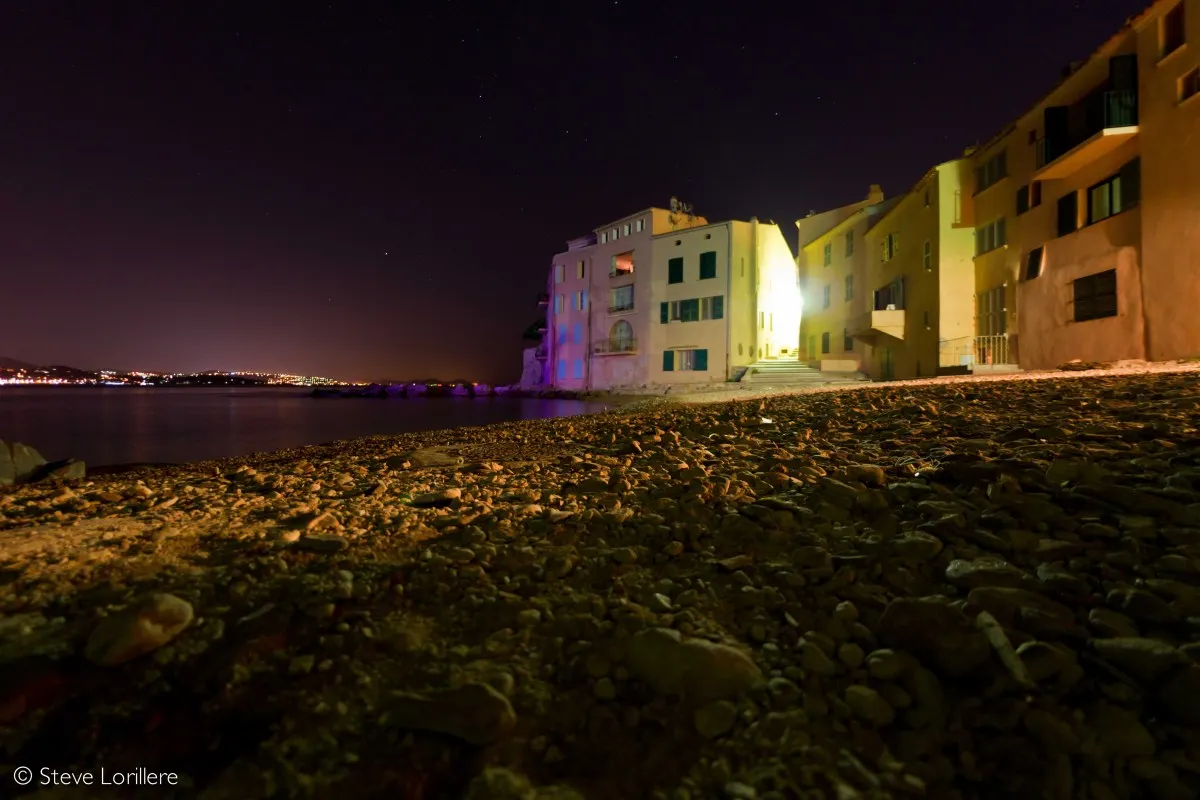 Night view of La Ponche's pastel waterfront buildings in Saint-Tropez, lit dramatically against a starry sky, pebble beach in foreground.