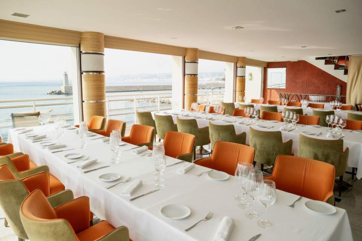 Dining room with orange and green chairs, white tablecloths, and large windows showing the sea and harbor
