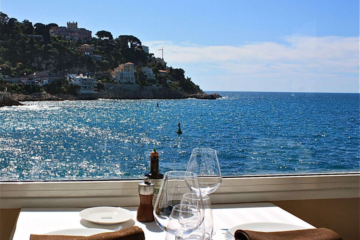 Table set with wine glasses by a window overlooking a rocky coastline and turquoise sea