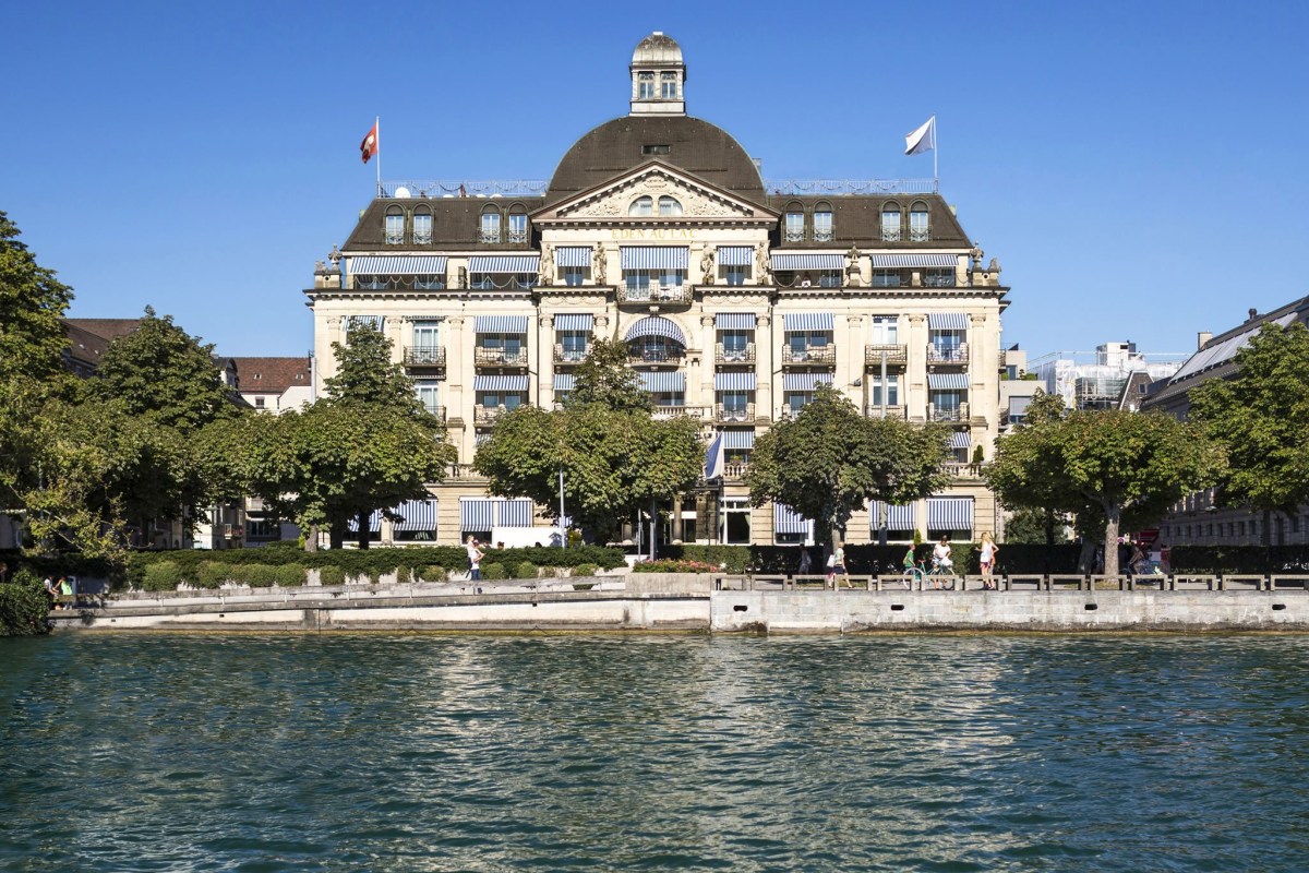 Waterfront view of La Réserve Eden au Lac hotel's grand façade with striped awnings, seen across the lake with trees lining the shore.