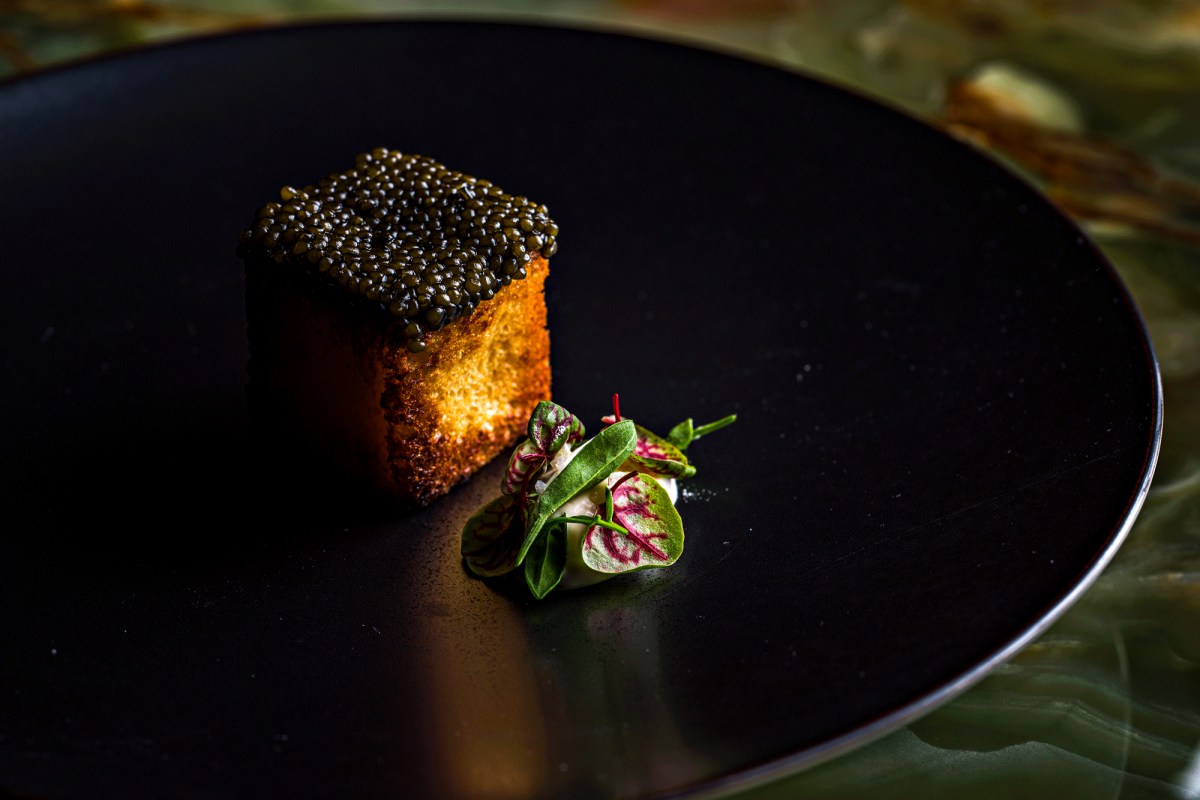Black plate with a caviar-topped brioche cube beside a small salad garnish
