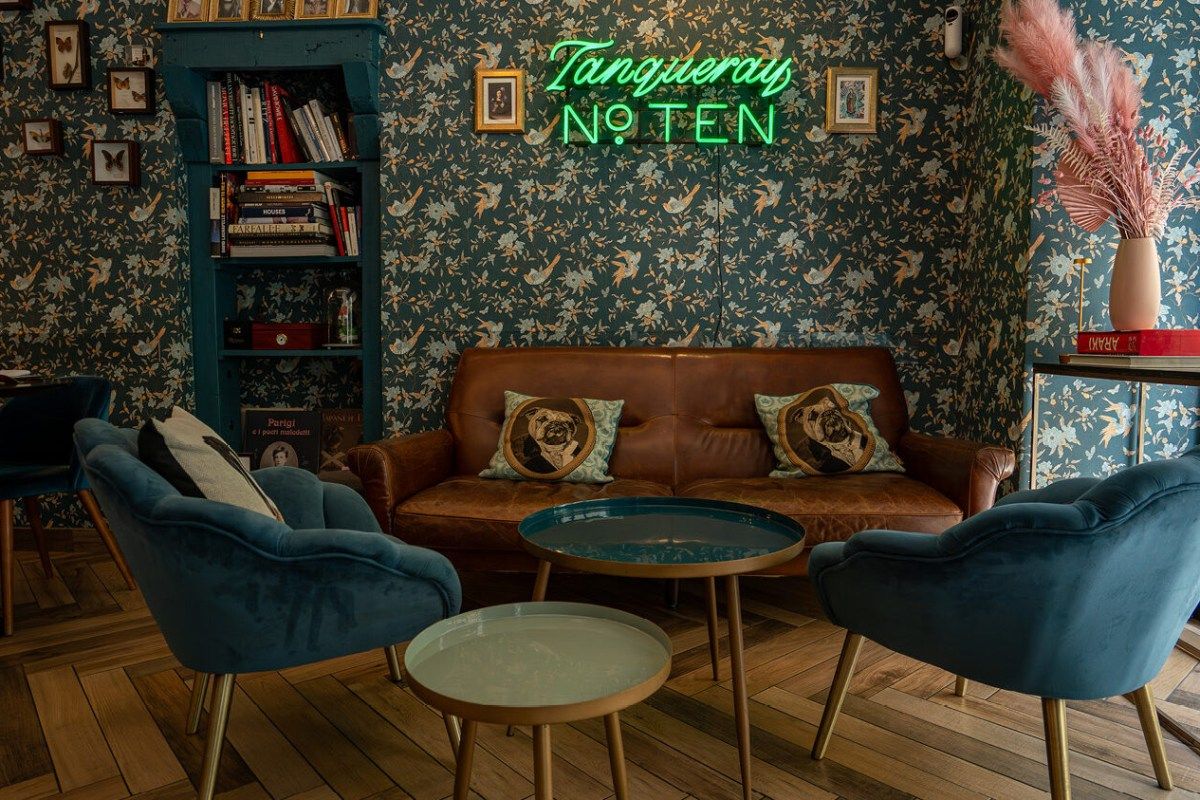 Lounge area with teal floral wallpaper, brown leather sofa, blue velvet armchairs, and a green Tanqueray neon sign