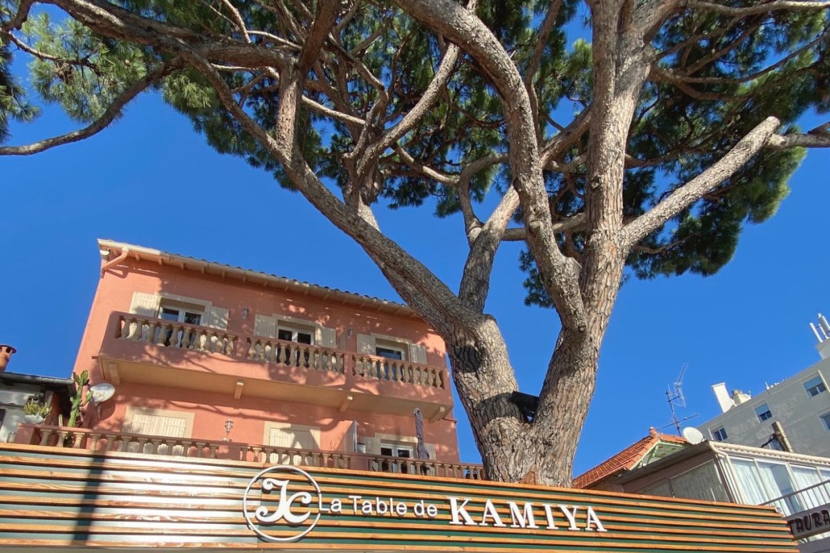 Exterior sign reading 'La Table de Kamiya' beneath a pine tree against a terracotta building and blue sky