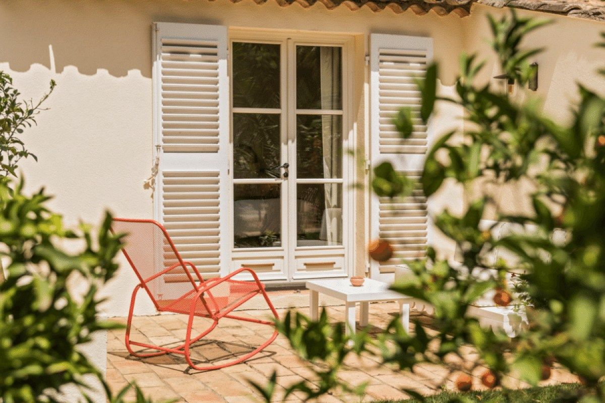 Exterior of a guest room with white shutters, French doors, terrace and orange chair framed by greenery