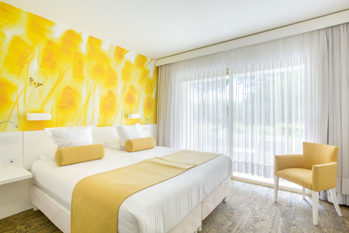 Hotel guest room with yellow wallpaper, bed, armchair, and sheer curtains by window