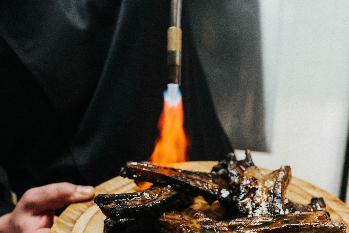 Chef torches a platter of glazed ribs with a blowtorch, flames visible
