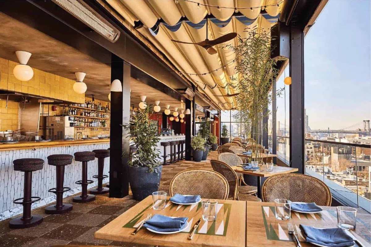 Rooftop dining room and bar with wicker chairs, string lights and a city skyline view through glass walls.