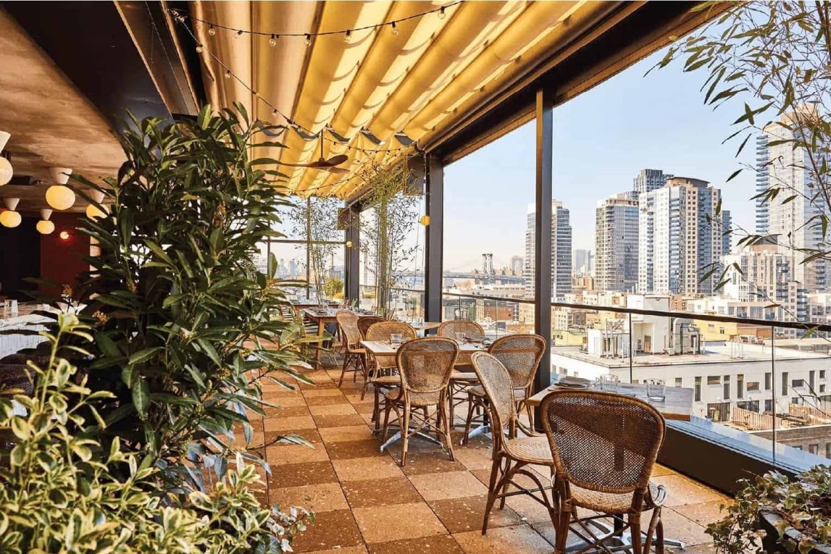 Open-air terrace with wicker chairs, string lights, bamboo plants and a sweeping Brooklyn skyline view.
