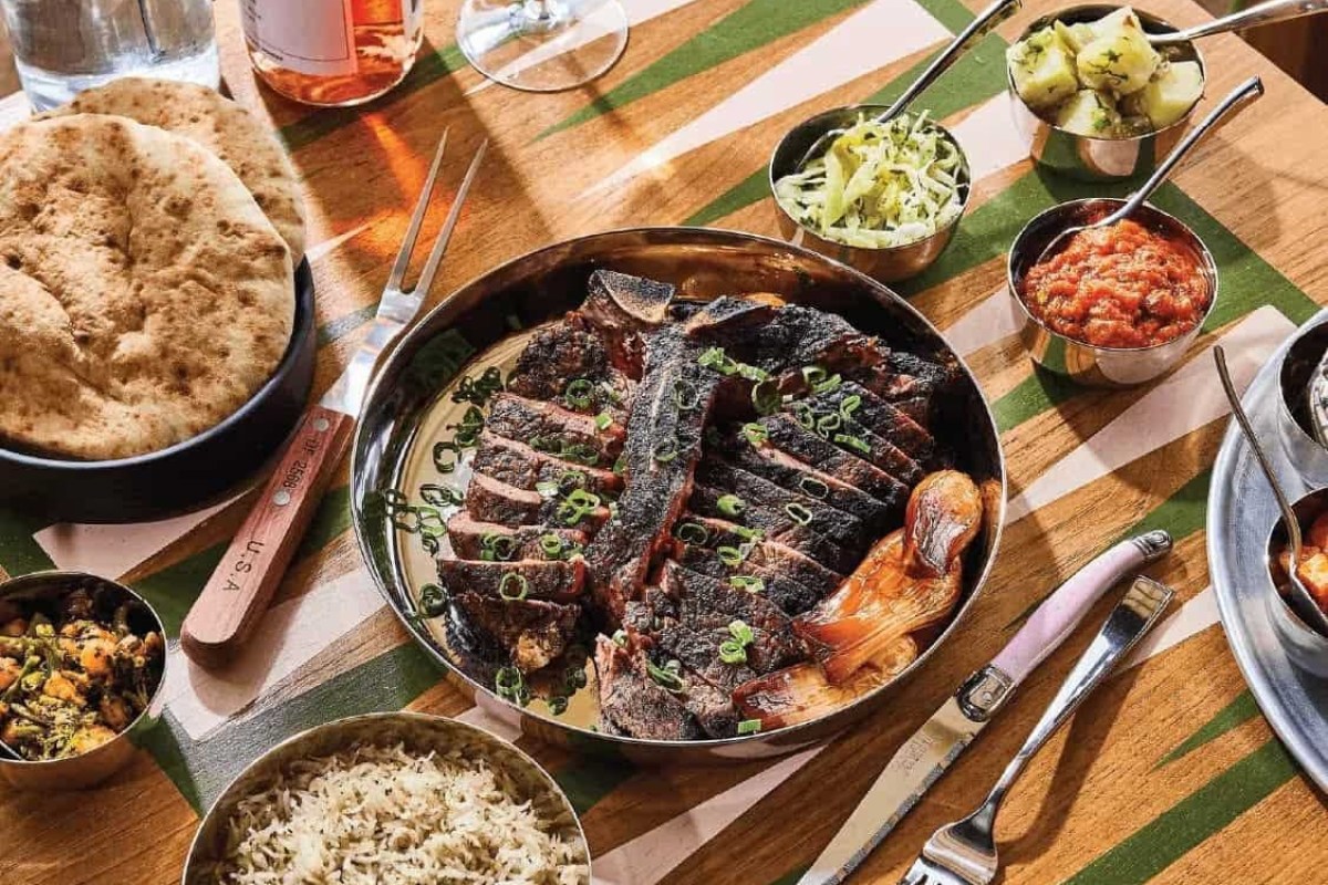 Laid table with sliced grilled steak, pita, rice, slaw and dips on a green-striped tablecloth.