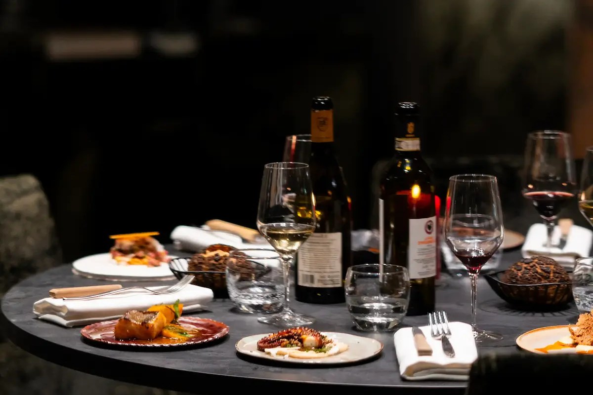 Dark restaurant table set with wine bottles, glasses, and several plated dishes