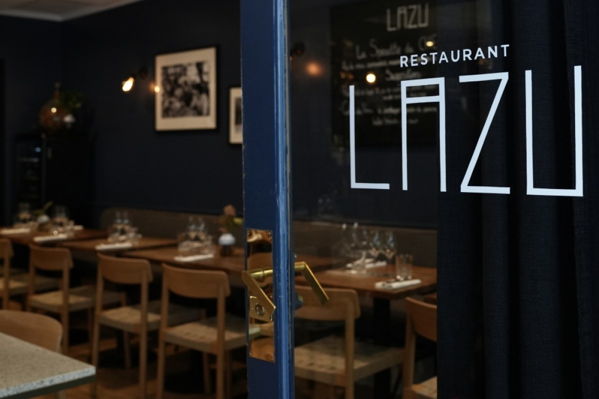 Entrance door of LAZU restaurant with glass signage, blue tones, dining room with set tables visible inside