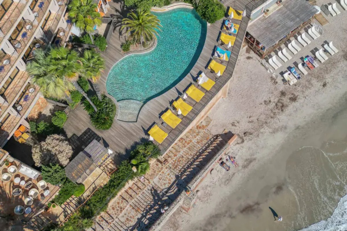 Aerial view of hotel building with curved pool, sun loungers, and beach beyond
