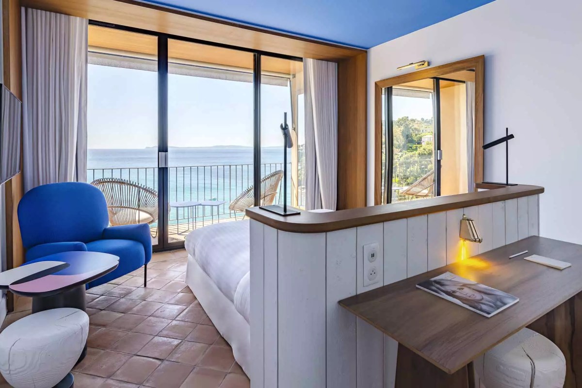 Modern hotel room with blue accents, bed, desk, and balcony overlooking the sea