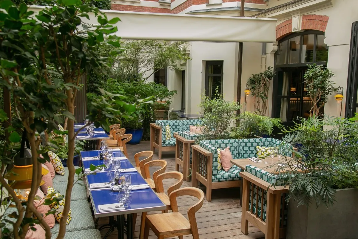 Outdoor courtyard terrace with dining tables, greenery and hotel facade with brick arches visible