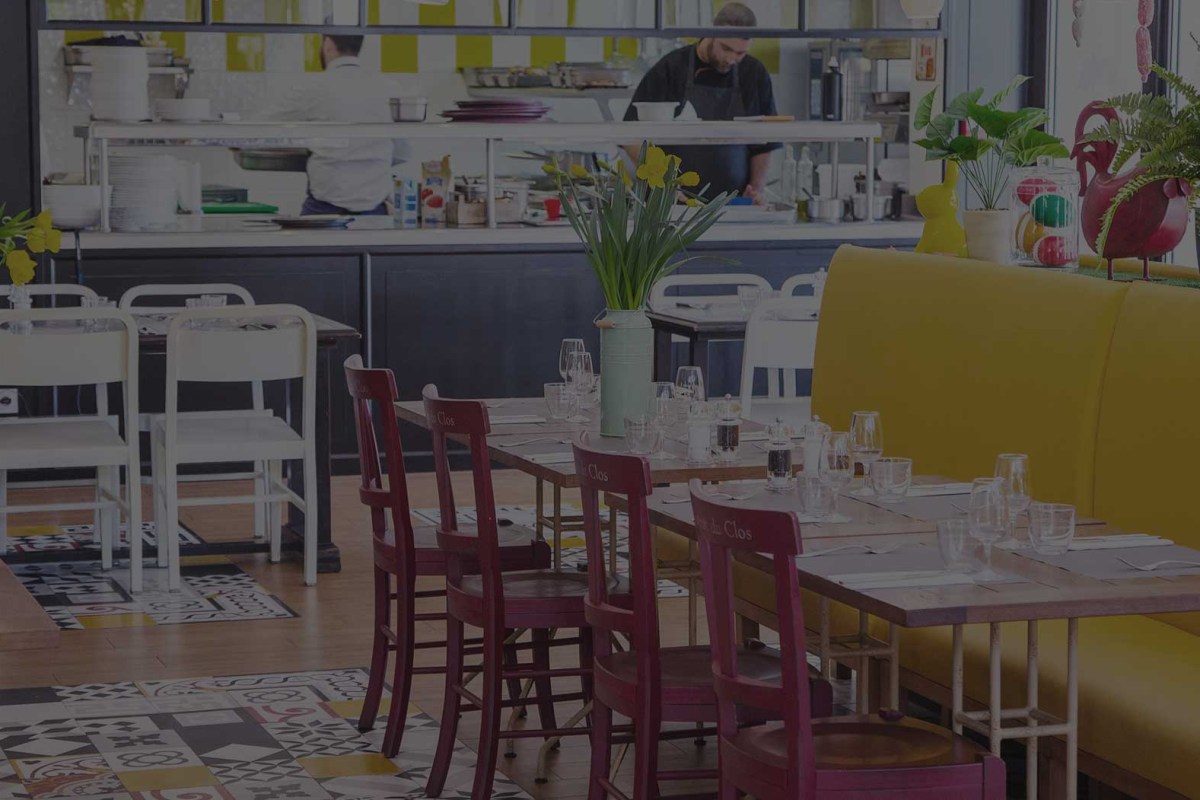Colorful dining room with yellow banquette, red chairs, set tables with glasses, chefs visible in open kitchen behind.