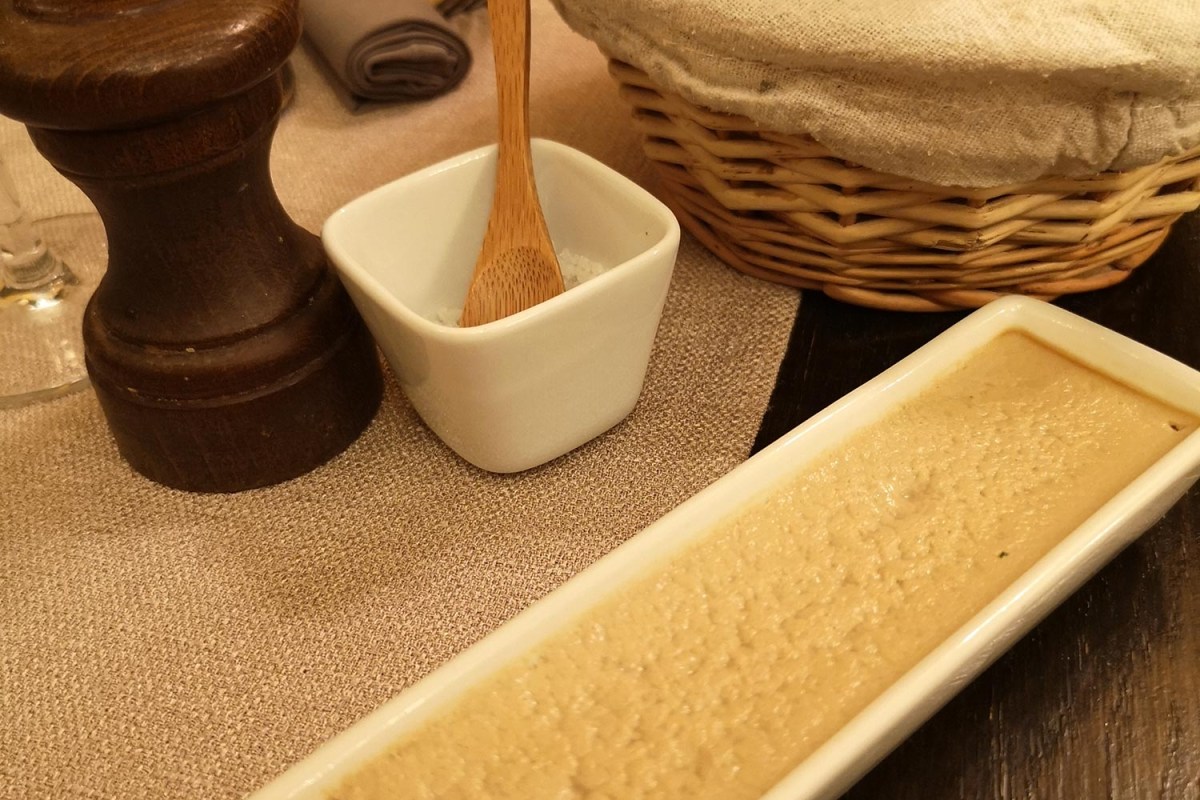 Close-up of a pâté terrine on a plate with a small salt dish, wooden spoon, and bread basket on the table