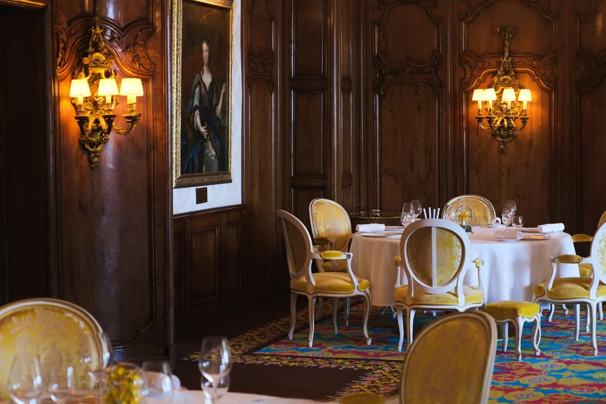 The dining room of Le Chantecler at Le Negresco in Nice, with carved wood panelling, an 18th-century portrait, wall sconces and gilded chairs around white tables