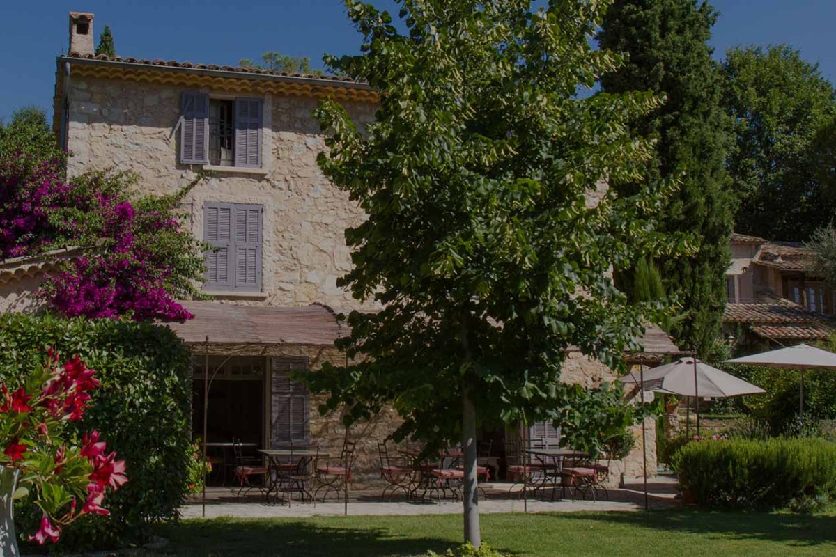 Provençal stone building exterior with lavender shutters, terrace tables, trees and flowering shrubs