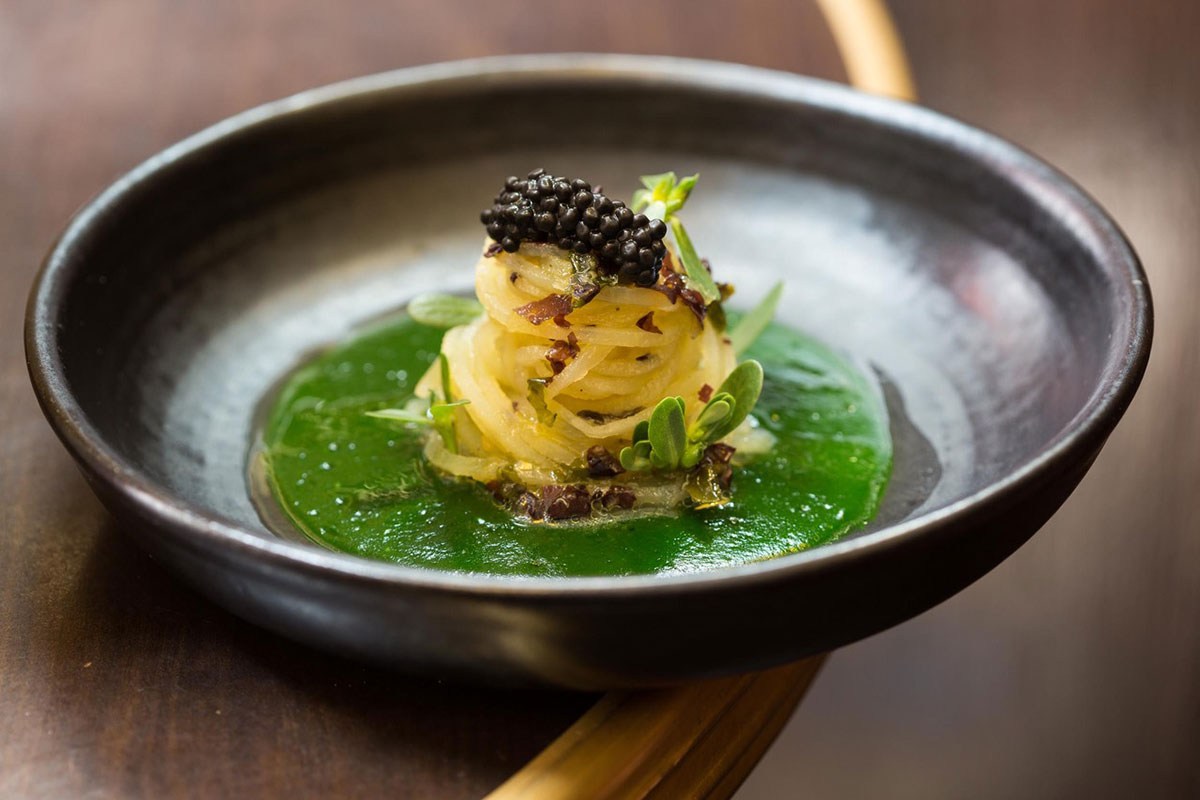 A plated dish: julienned vegetable nest topped with caviar on a green sauce base, served in a dark bowl.