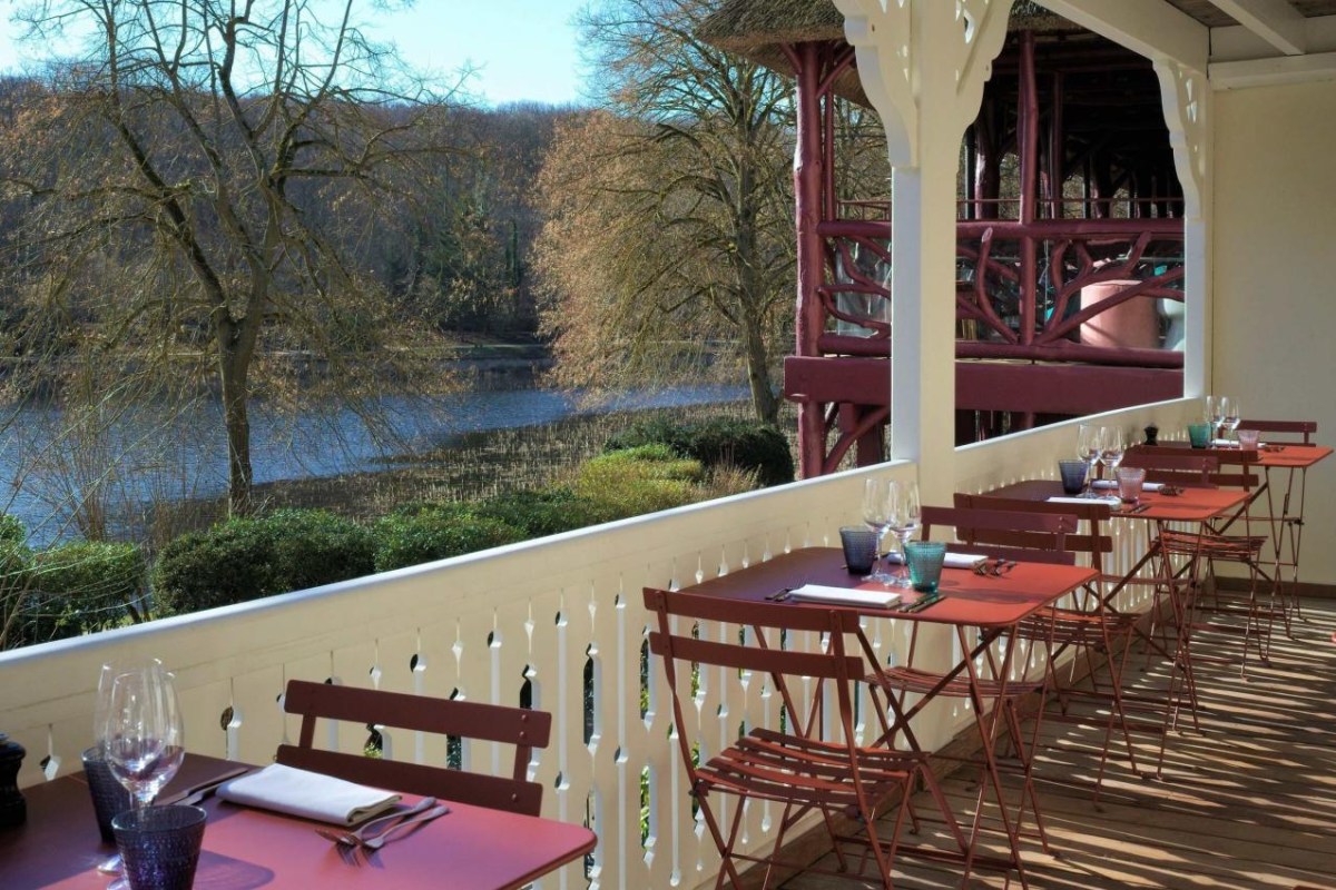Restaurant terrace with red bistro tables set for dining, overlooking a lake and trees