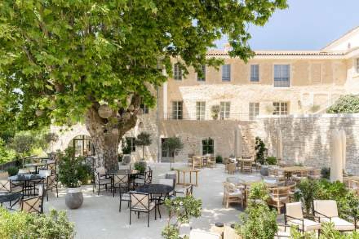 Stone hotel façade with shuttered windows seen across a sunny courtyard terrace with outdoor dining tables under a large tree
