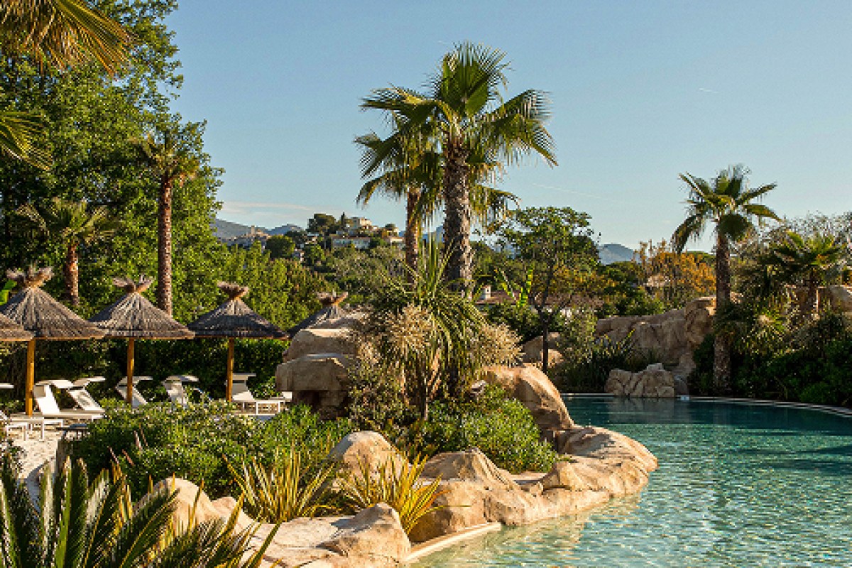 Resort pool with rocks, palm trees and thatched umbrellas, hillside village visible in background