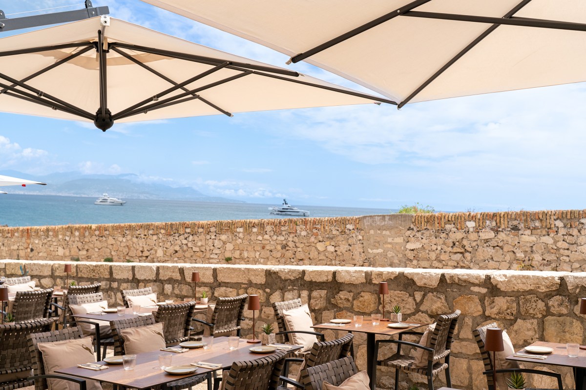 Seaside terrace with cushioned chairs, wooden tables set for dining, stone wall and sea view with yachts in the background