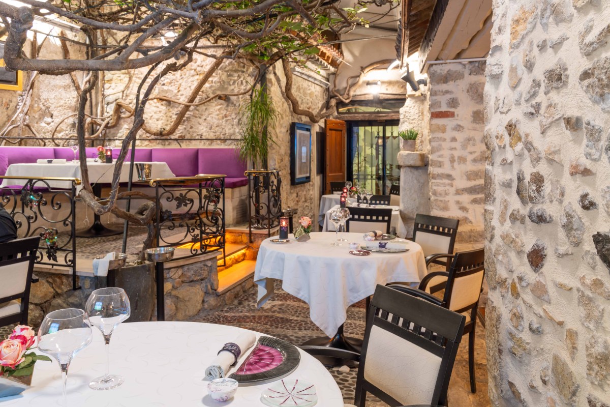 Elegant stone-walled dining terrace with vine canopy, purple banquette and tables set with white linens and wine glasses
