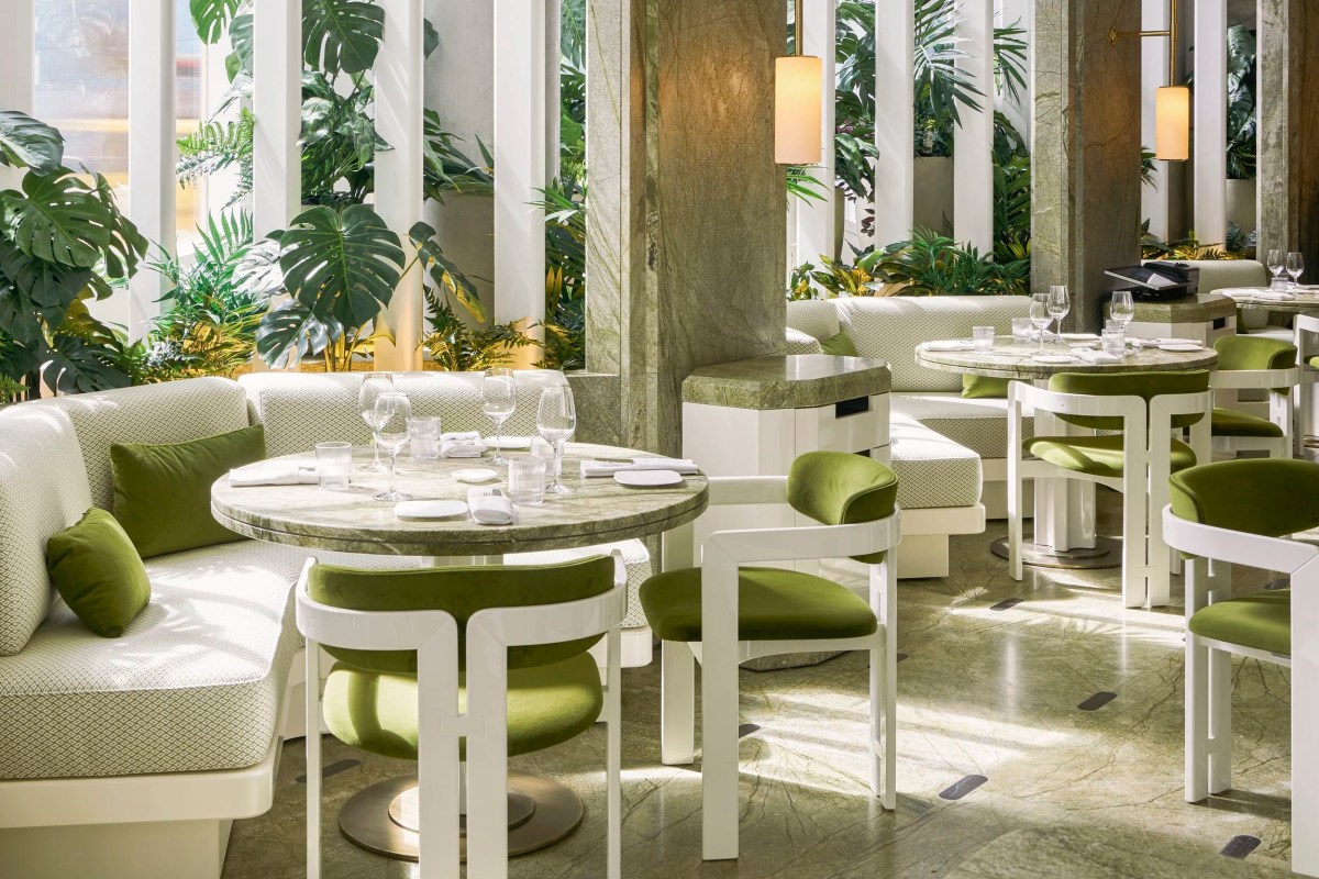 Elegant dining room with marble tables, green velvet chairs, and lush greenery between white columns