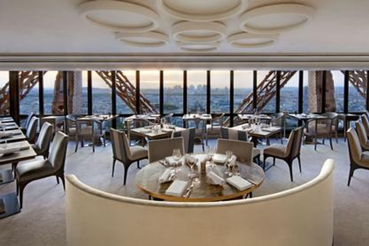 Interior dining room of Le Jules Verne with tables set for service and a panoramic view of Paris through Eiffel Tower ironwork.