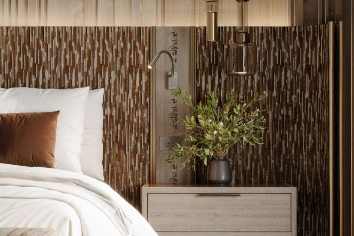 Close-up of a hotel bed with white linens, a textured wood headboard, bedside lamp, and a vase of greenery.