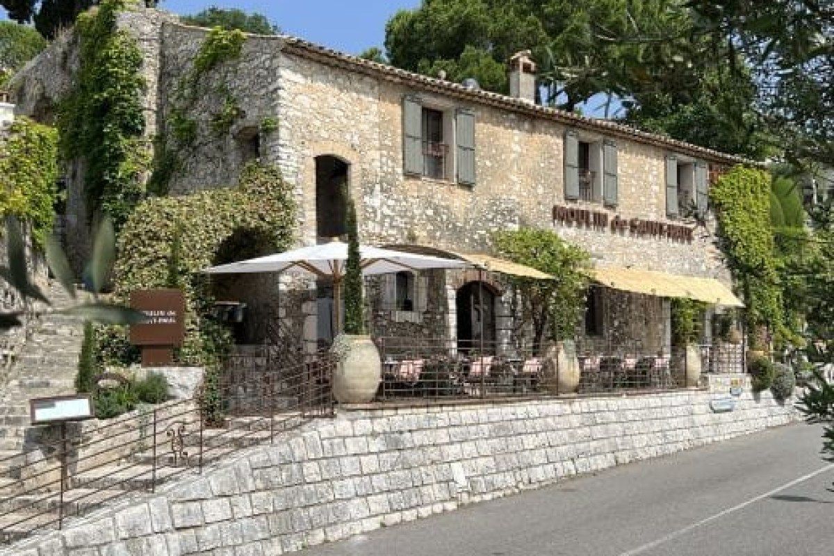 Stone exterior of Moulin de Saint-Paul restaurant with vine-covered facade, terrace seating and umbrella under trees