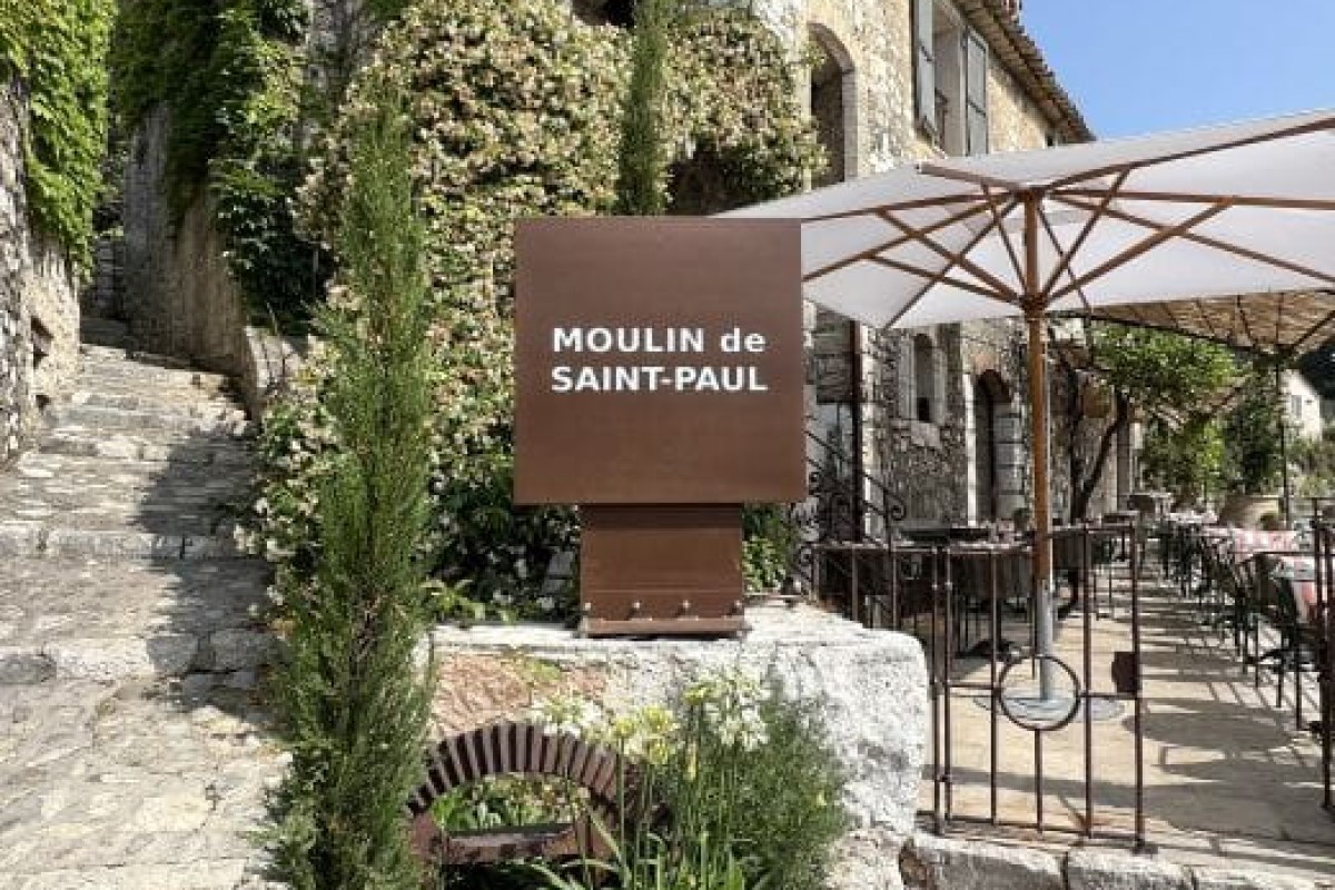 Entrance sign reading 'Moulin de Saint-Paul' beside stone steps with terrace tables and umbrellas visible