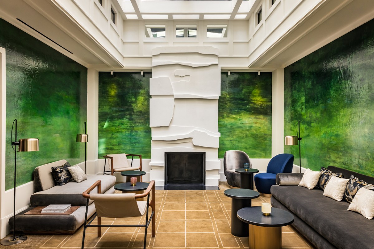 Hotel lounge with green mural walls, modern seating around a white stone fireplace under a skylight