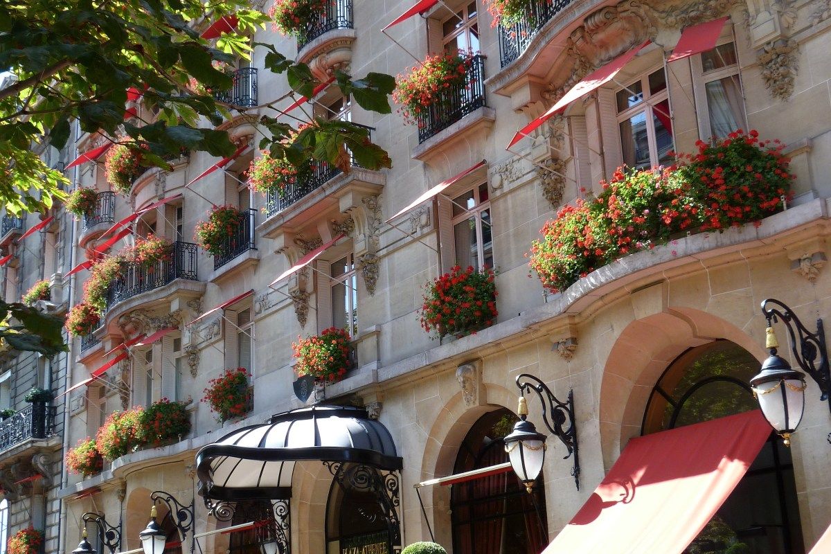 Facade of Plaza Athénée Paris with red awnings, flower-filled balconies, ornate stone details and entrance lanterns