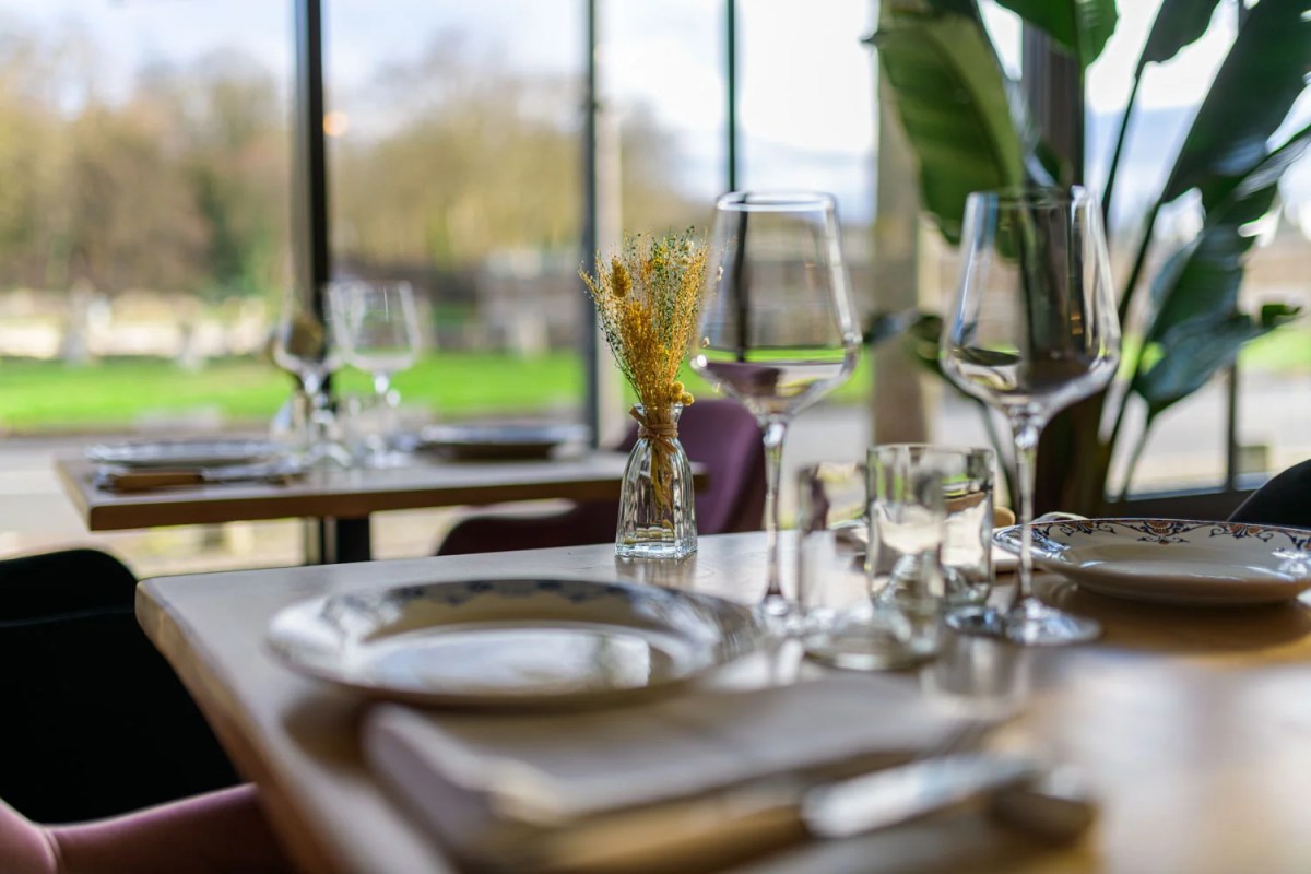 Restaurant dining room with set tables, wine glasses, and a window view onto a green park.