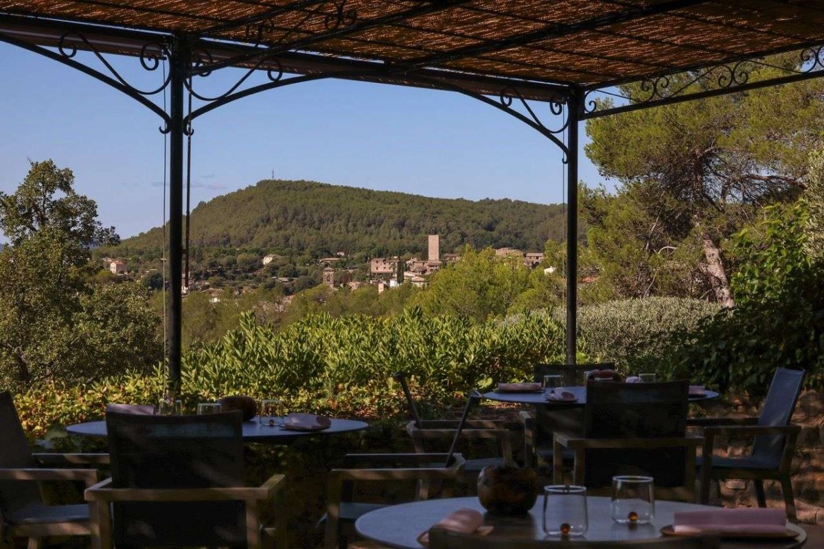Outdoor terrace under a pergola with set tables, overlooking hills and a hilltop village.
