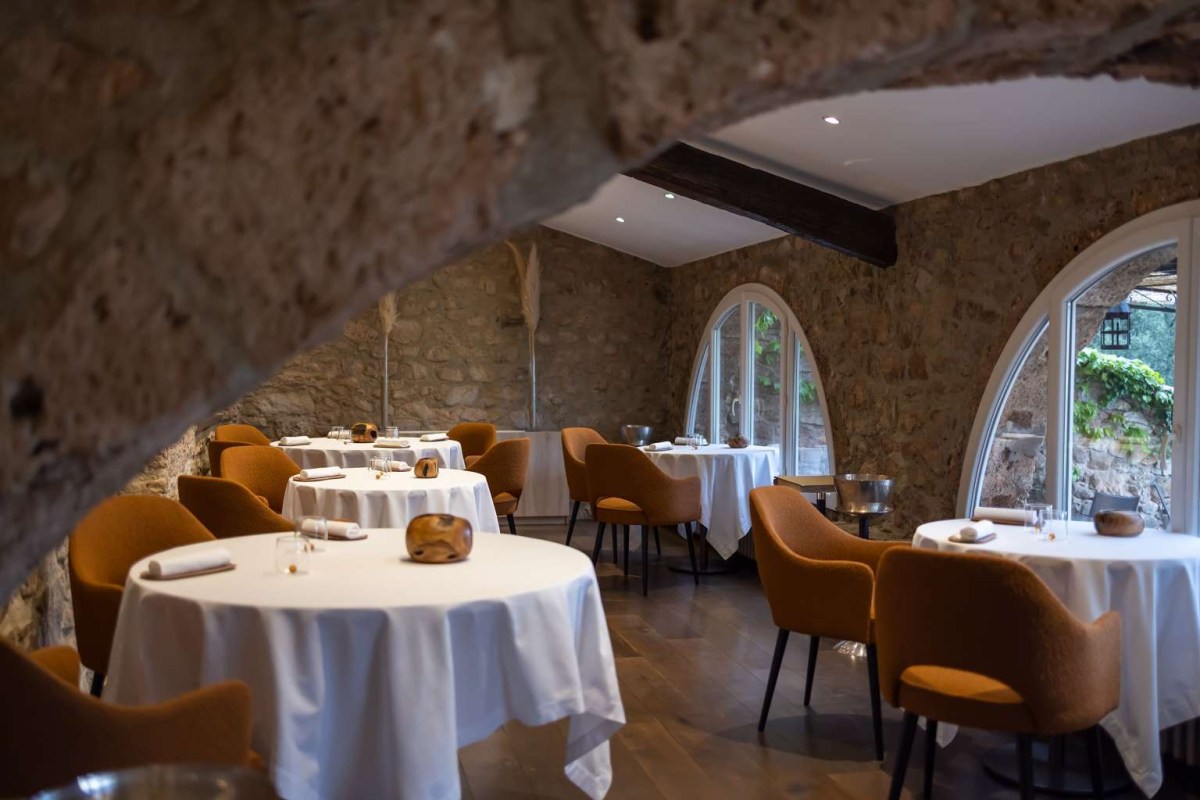 Elegant stone-walled dining room with white-clothed round tables and orange chairs, arched windows to garden.