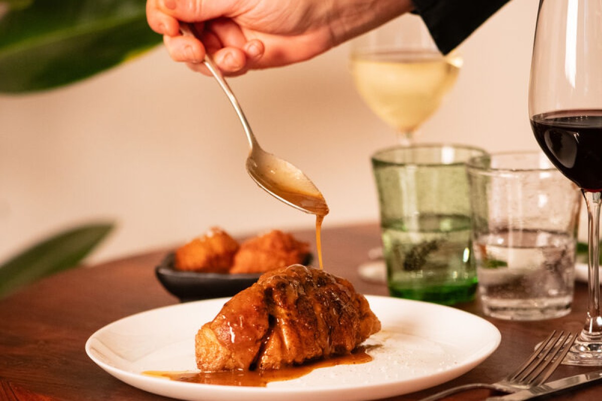 Hand spooning sauce over a plated meat dish with wine glasses on table at Le Socle