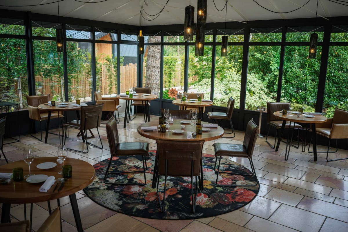 Glass-walled dining room with round tables set for dinner, garden view and floral rug