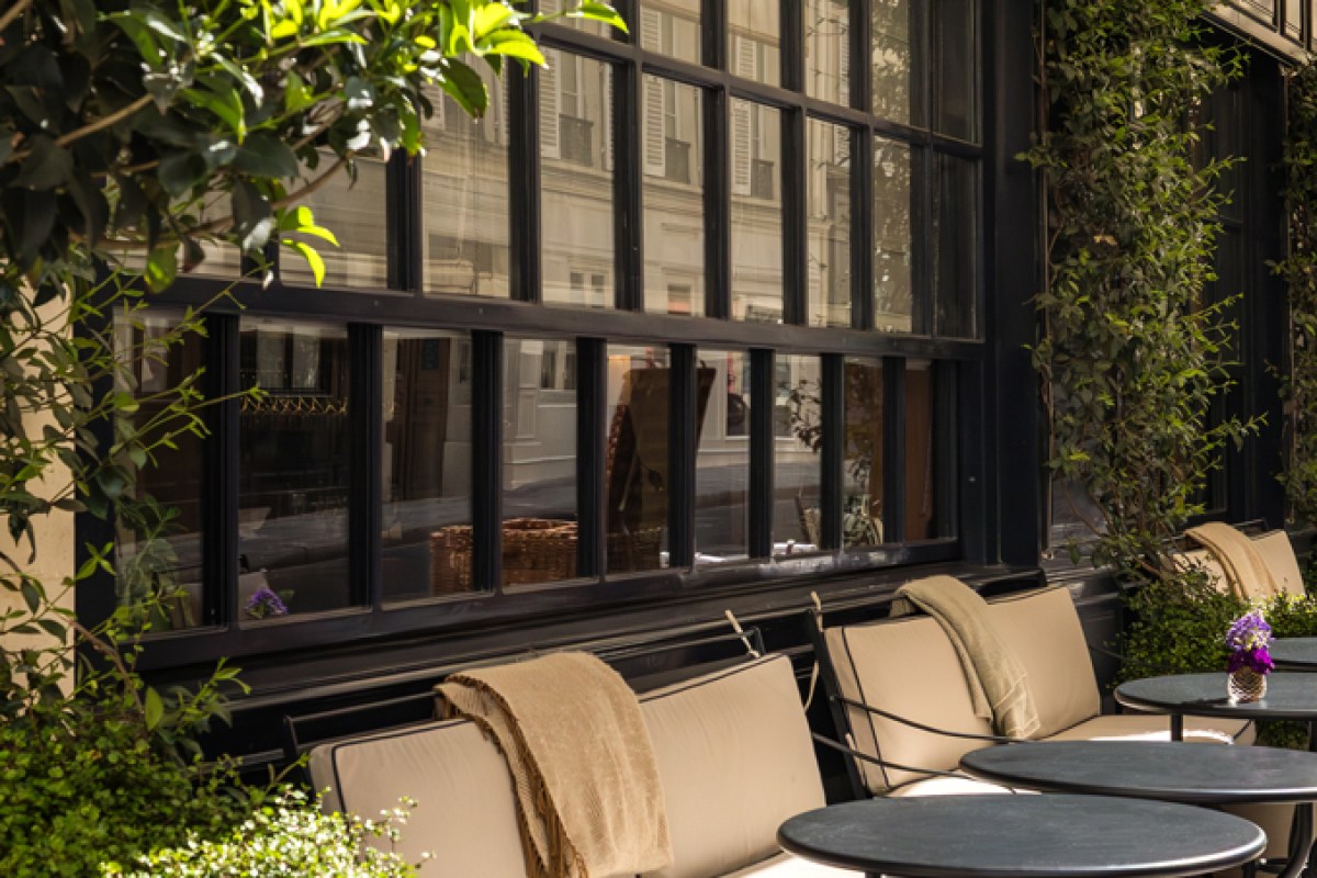 Restaurant exterior terrace with cushioned chairs, dark window frames, greenery and small tables outside