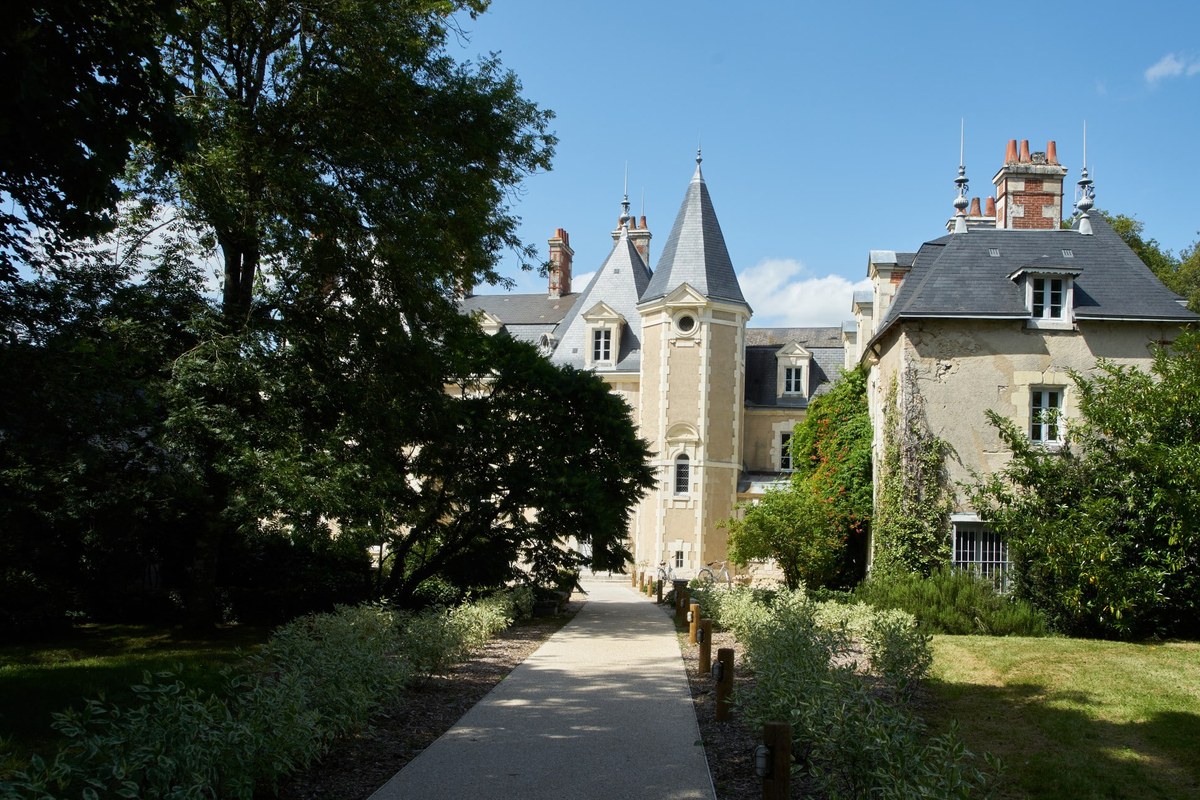 Château-style building with turrets and slate roofs seen along a garden path lined with trees and shrubs