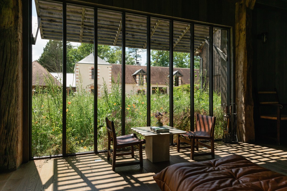 Interior lounge nook with leather chairs and table framed by large windows overlooking a wildflower garden and outbuildings